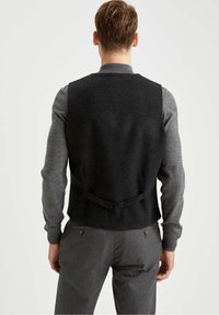 Schwarze Wollweste mit glatter Textur, ausgestattet mit einer Schnalle auf dem Rücken. Das Modell trägt ein graues Langarmshirt mit gerippten Bündchen und graue Hosen.