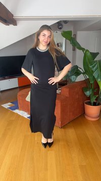 Young woman in a long black dress with pearl neckline and black heels stands with hands on hips in a modern living room.