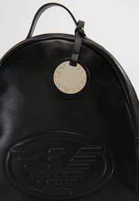 Sac à dos en cuir noir à forme arrondie, avec un logo en relief bien visible et une étiquette circulaire de la marque en métal argenté. Texture lisse.