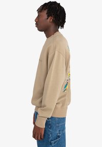 Sudadera color crema con un corte relajado, que presenta un vibrante bordado de pájaro en el lado izquierdo y puños y dobladillo acanalados.