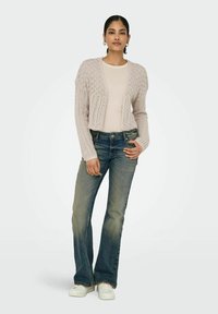 Cardigan a maglia rosa chiaro con apertura frontale, indossato sopra un top aderente color crema, abbinato a jeans di denim svasati e sneakers bianche.