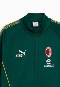 Zelená športová bunda s celým zipsom, vysokým golierom, žltými detailami, logom Puma, nápisom "KING" a emblémom ACM 1899 na hrudi.