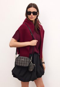 Černá a zlaté vzorovaná krosbody taška s odpovídající malou kapsičkou, obě s zlatými detaily a černým popruhem, nošené proti bordó outfitu.