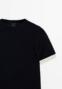 Haut en maille noir à manches courtes avec motif ajouré texturé et bordure côtelée blanche aux extrémités des manches sur fond blanc uni.