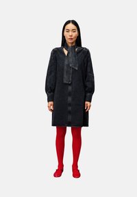 Vrouw met lang zwart haar, gekleed in een donkere spijkerjurk met een strik om de nek, helderrode panty’s en glanzende rode schoenen, staand tegen een witte achtergrond.