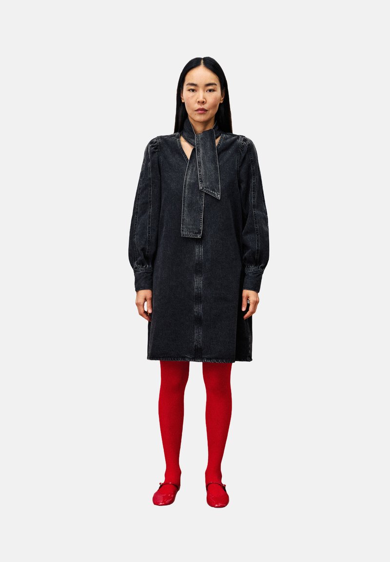 Vrouw met lang zwart haar, gekleed in een donkere spijkerjurk met een strik om de nek, helderrode panty’s en glanzende rode schoenen, staand tegen een witte achtergrond.