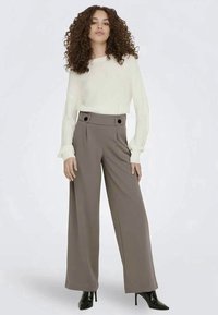 Krémový pletený svetr a široké kalhoty světle hnědé barvy s knoflíkovými detaily, doplněné černými lodičkami na vysokém podpatku. Jednoduchý, texturovaný design.