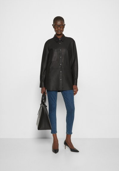 Camisa de couro preta com gola, fecho de botão e bolso frontal; combinada com jeans skinny azuis e sapatos de salto agudo pretos.
