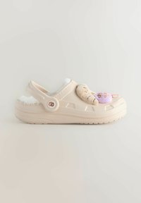 Sabots beiges avec doublure en fourrure, ornés d'un ours en peluche et d'un donut décoratifs sur le dessus. Forme arrondie et bride arrière ajustable. Semelle texturée.