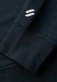 Marine sweatshirtmanschet med en struktureret stof, der har en hvid logo-detalje. Glat, tyk vævning er synlig, hvilket fremhæver holdbarhed og komfort.
