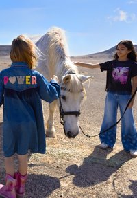 Deux enfants interagissent avec un cheval blanc. L'un porte une veste en denim avec des lettres colorées "POWER" ; l'autre porte un tee-shirt graphique noir et un jean large.