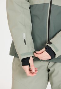 Grüne wasserdichte Jacke mit strukturierter Oberfläche, mit einem verstellbaren Klettverschluss am Ärmel und schwarer Innenauskleidung. Markenlogo auf dem Ärmel sichtbar.