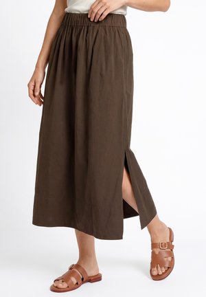 Vrouw met een bruine midi-rok met zijsplit, gecombineerd met leren platte sandalen in tan met gespsluiting.