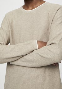 Pull beige texturé avec un col rond et un bord blanc contrastant au niveau du col et des poignets, doté de manches longues et d'une coupe ample.