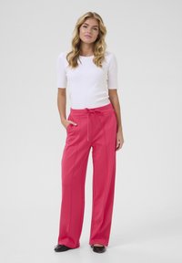 Pantalon large rose avec taille à cordon, doté de poches latérales et de coutures verticales ; associé à un haut blanc ajusté.