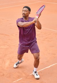 Ljubičasta atletička majica i šorts na muškom igraču koji zamahuje teniskim reketom na zemljanom terenu, nosi bijele čarape i crne teniske cipele.