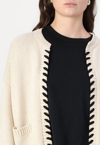 Beige gestrickte Strickjacke mit einem strukturierten Rand, verziert mit schwarzen Schnürsenkeln und einer Tasche, getragen über einem schwarzen T-Shirt. Nahaufnahme.