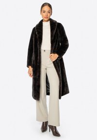 Bruine nepbont lange jas met een brede kraag, gecombineerd met een crèmekleurig top en lichtgrijze wijde broek. De textuur lijkt zacht en pluizig.