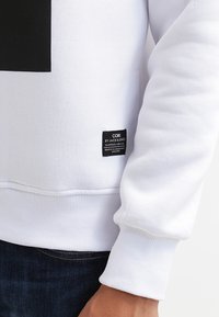 Weißer Pullover mit geripptem Saum und Bündchen; verfügt über ein schwarzes quadratisches Design auf der Brust und ein Stoffetikett an der Seite.