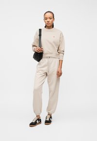 Γυναίκα με πλεξούδες φοράει μπεζ φούτερ και φόρμες, μαύρη τσάντα χιαστί και μαύρα-άσπρα αθλητικά παπούτσια, στέκεται μπροστά σε λευκό φόντο.