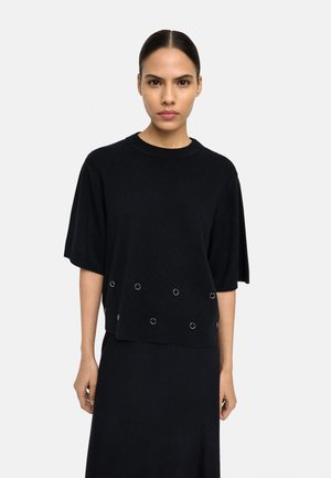 Kobieta z zaczesanymi do tyłu włosami, ubrana w czarną bluzkę z krótkim rękawem z srebrnymi oczkami przy krawędzi oraz dopasowaną czarną spódnicę.