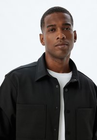 Chaqueta de camisa de algodón negra con un cierre de botones en la parte delantera, dos bolsillos frontales y un cuello clásico. Camisa interior blanca y nítida visible debajo.