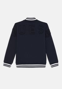 Giubbotto bomber blu navy con polsini e colletto a righe bianche. Caratterizzato dal testo in rilievo "GUESS" sul retro. Tessuto morbido e vestibilità comoda.