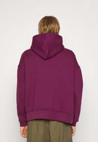Paars oversized hoodie met een grote platte capuchon, ribbelboord onderaan en aan de mouwen, gemaakt van zachte stof, gezien van achteren.