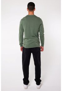 Grüner Strickpullover mit langen Ärmeln, Rundhalsausschnitt und körpernahem Saum, kombiniert mit schwarzen Jeans und weißen Turnschuhen. Strukturierter Stoff sichtbar.