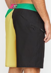 Shorts de bain noirs et jaunes avec une ceinture vert et rose, comportant une poche zippée sur le côté et des panneaux de couleur contrastée.
