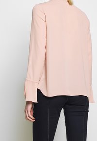 Blouse rose clair à manches longues, avec poignets plissés et ourlet arrondi. Confectionnée dans un tissu lisse, offrant un design minimaliste.