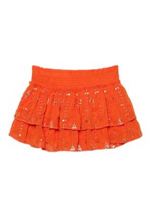 Femme aux cheveux mouillés portant un haut de bikini orange, une mini-jupe à volants assortie et des sandales à plateforme noires, debout devant un fond marron.