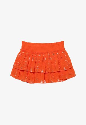 Kobieta z mokrymi włosami, ubrana w pomarańczowy top bikini, dopasowaną warstwową mini spódnicę i czarne sandały na platformie, stojąca na tle brązowego tła.