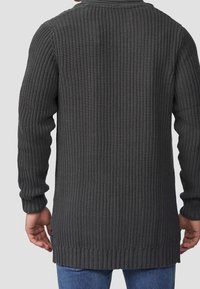 Hombre vistiendo un suéter de punto texturizado gris oscuro con puños y dobladillo acanalados, visto desde atrás, combinado con jeans azules.