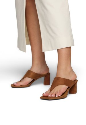 Voeten met bruine suède sandalen met blokhak en teenbandjes, gecombineerd met beige wijde broekspijpen met zijsplitten.