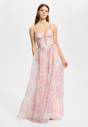 Jonge vrouw in een doorschijnende, mouwloze maxi-jurk met pastelkleurige bloemenprint, diepe V-hals en dunne bandjes, staand tegen een effen achtergrond.