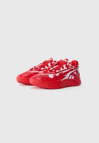 Par af røde og hvide atletiske sneakers med dristige mønstre og tykke såler mod en ensfarvet lys grå baggrund.