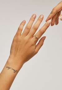 Anillo de caballito de mar de oro en el dedo anular de una mano de tono de piel medio; uñas al natural, tatuaje visible en la muñeca.