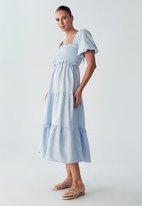 Svetlo modra maksi obleka s nabranim korzetom, napihnjenimi rokavi in večplastnimi volančki. V kombinaciji z svetlo obarvanimi ravnimi sandalami. Gladka tekstura tkanine.