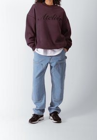 Lilla overdimensioneret sweatshirt med "Atelier" i sort skrift, parret med lyseblå posebukser og sorte sneakers med hvide såler.