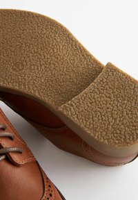 Chaussure en cuir brun avec une semelle en caoutchouc texturé. La semelle présente une surface à motifs, et la chaussure dispose de coutures décoratives et de fermetures à lacets.