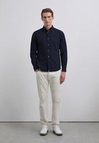 Chemise à boutons bleu marine avec poche poitrine, associée à un pantalon couleur crème et des baskets blanches. Design minimaliste et texture de tissu lisse.