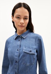 Jeanshemd mit Knopfverschluss vorne, klassischem Kragen, langen Ärmeln und einer Brusttasche. Die Textur ist leicht ausgewaschenes Blau.