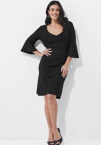 Schwarzes Kleid mit V-Ausschnitt, Glockenärmeln und seitlichen Schlitzen, aus glattem Stoff, kombiniert mit schwarzen offenen High Heels.