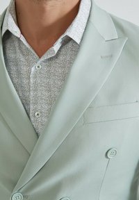 Blazer vert menthe clair avec un design à double boutonnage, associé à une chemise à col avec une base blanche et un motif floral vert.