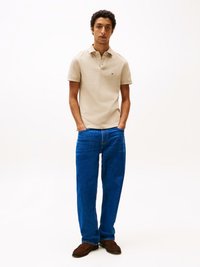 Beige színű rövid ujjú póló, egygombos gallérral és logóval. Kék egyenes szárú farmer és barna slip-on cipő egészíti ki az öltözéket.