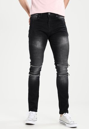 Jean noir slim avec texture côtelée sur les cuisses et détails zippés au-dessus des genoux, porté avec des baskets blanches.
