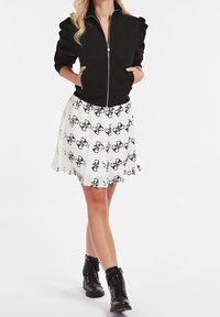 Chaqueta negra con cremallera y mangas abullonadas, combinada con una falda blanca plisada con patrones abstractos en negro. Completado con botas negras hasta el tobillo.