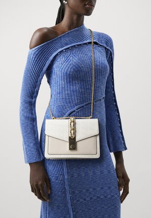 Vrouw in een blauwe asymmetrische geribbelde gebreide jurk met een witte leren crossbodytas met gouden ketting- en sluitingsdetails.