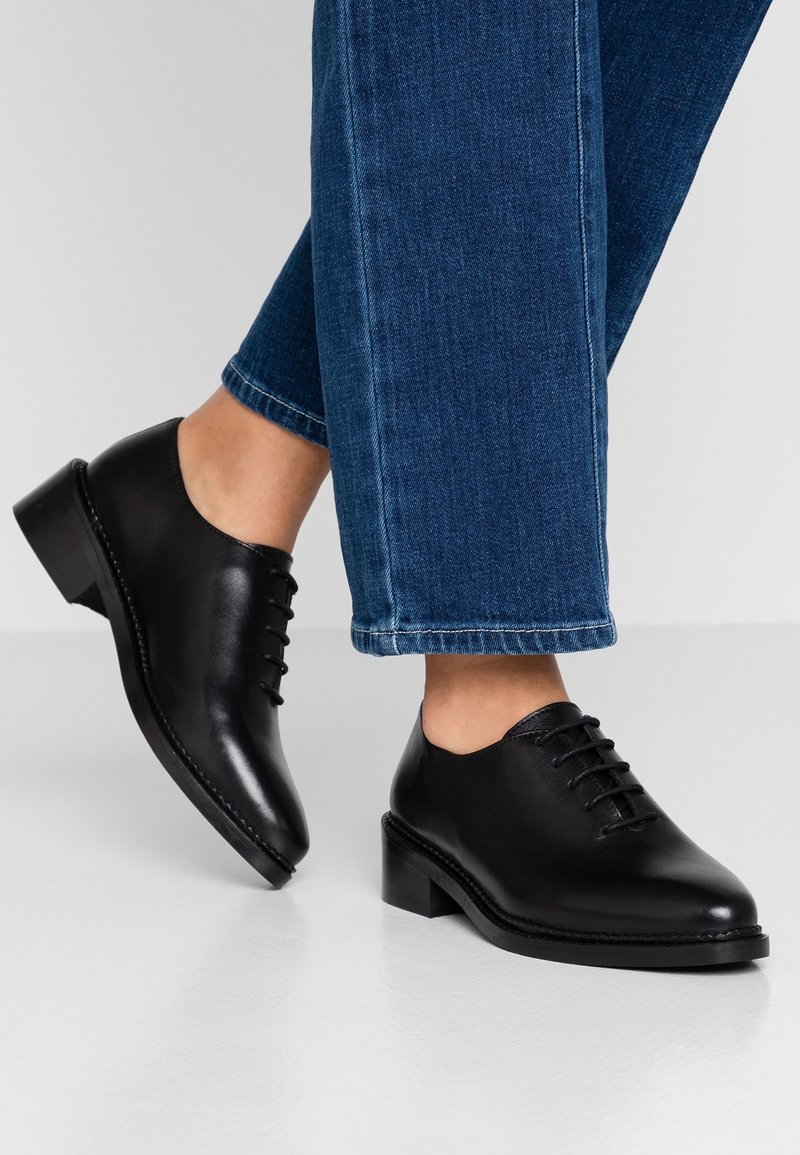 Chaussures noires en cuir à lacets, avec un bout arrondi, un talon bas, et des détails de couture, portées avec un jean bleu cropped.